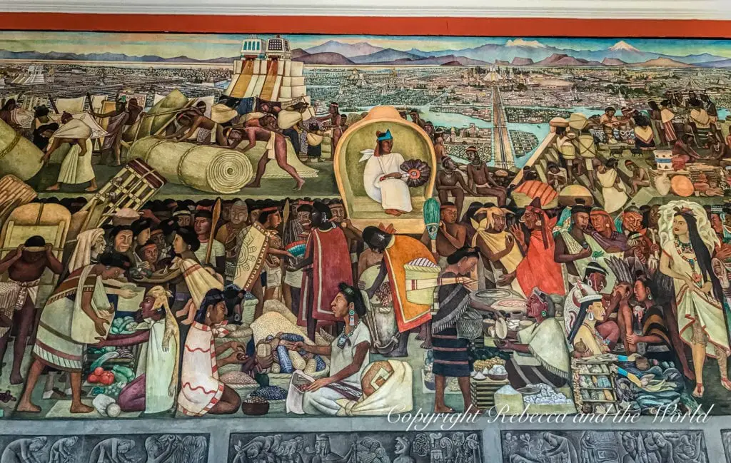 A vibrant Diego Rivera mural depicting a bustling market scene with various figures engaging in trade and carrying goods in front of a depiction of ancient Aztec pyramids and the cityscape. This mural is located in the Palacio Nacional in Mexico City.