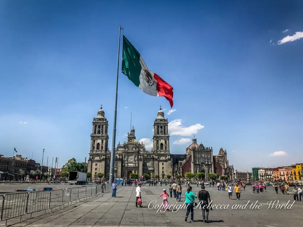 Mexico City's Zócalo with the towering Metropolitan Cathedral in the background, a large Mexican flag waving prominently in the foreground, and people milling about the square.