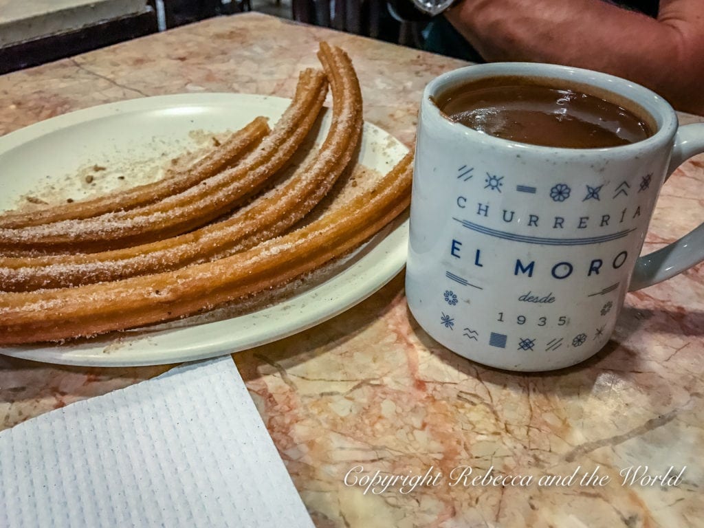 A plate of freshly made churros, dusted with sugar, next to a mug of hot chocolate, both resting on a marble cafe table.