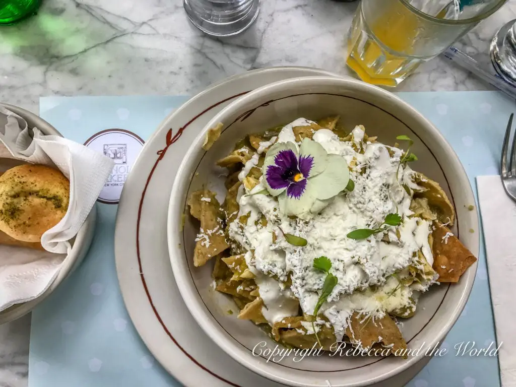 A dish of chilaquiles topped with white cream, crumbled cheese, and a delicate purple edible flower, served with a side of bread on a marble table. Chilaquiles are a delicious breakfast staple in Mexico City.