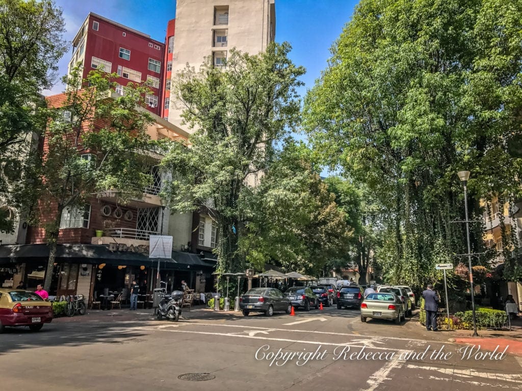 A tree-lined street in Mexico City's Condesa neighborhood, with outdoor cafes and brick buildings, bustling with pedestrians and parked cars on a sunny day.