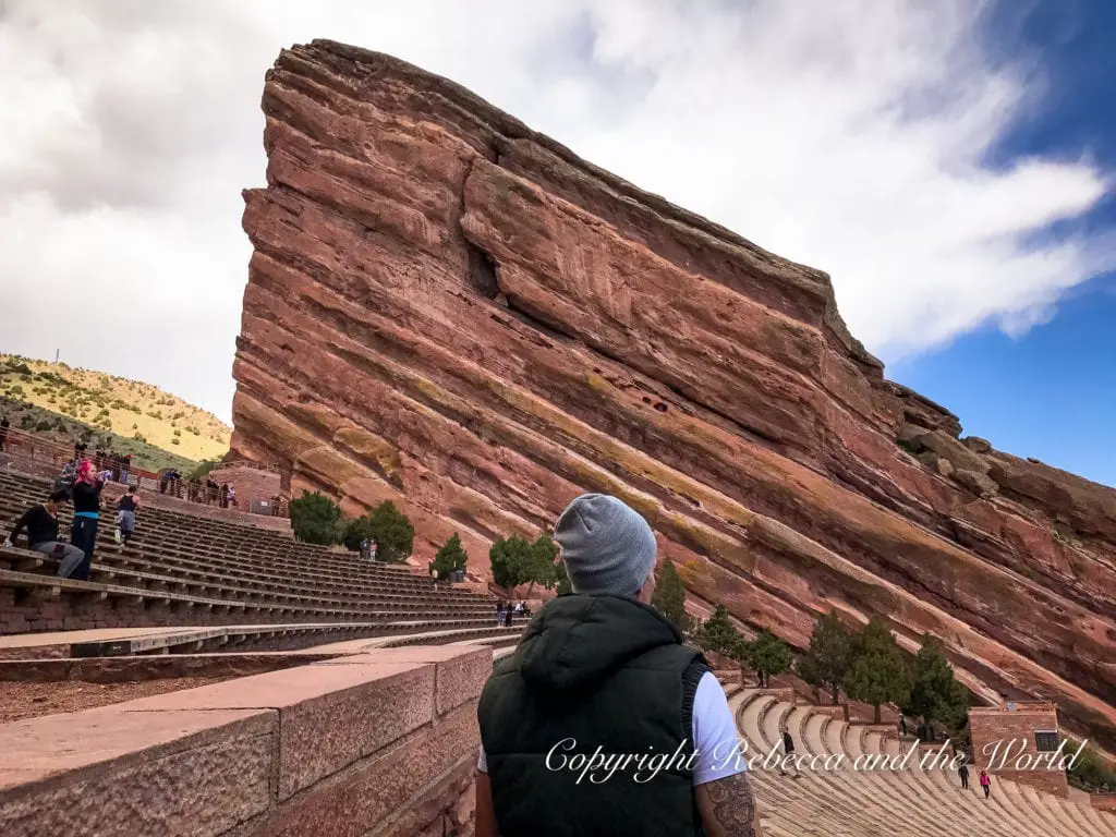 A person - the author's husband - in a grey beanie and black vest standing with their back to the camera, looking up at a towering red rock formation at Red Rocks Amphitheatre. The sky is partly cloudy.