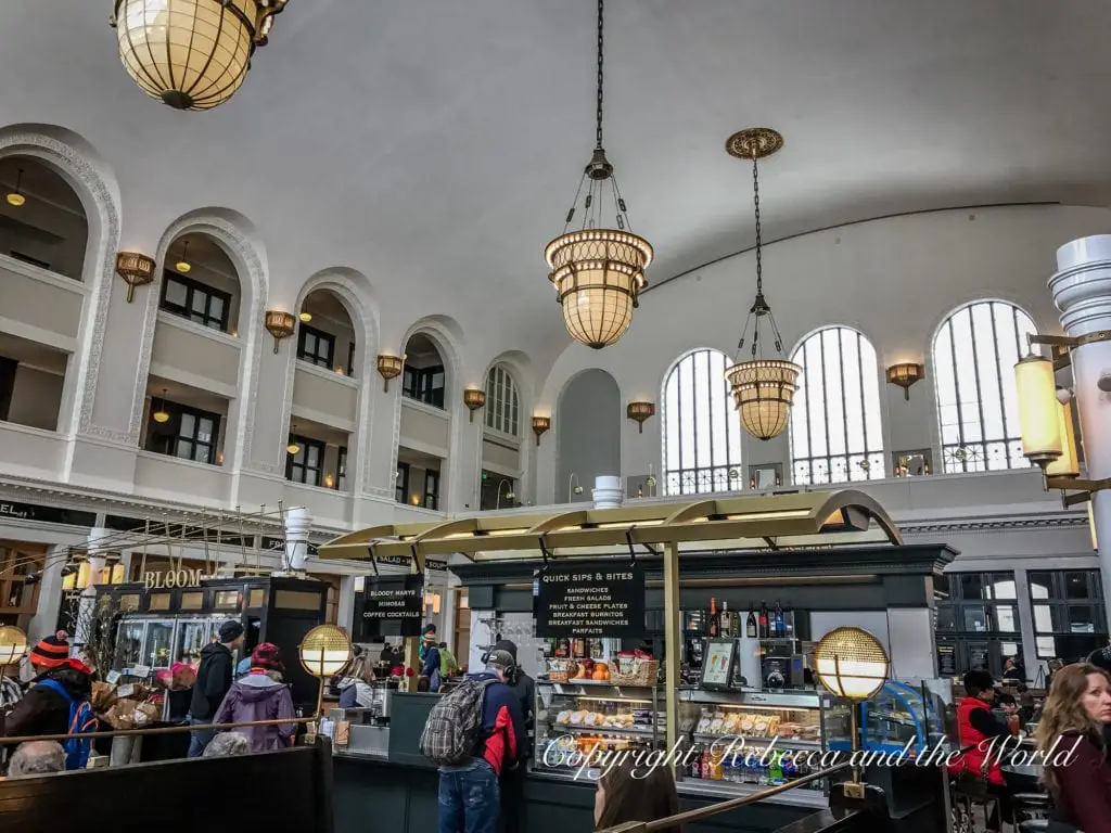 A busy indoor marketplace with high ceilings, arched windows, and elegant hanging lamps. Patrons are gathered at various food stalls. This is the inside of the beautiful Union Station in Denver, Colorado.