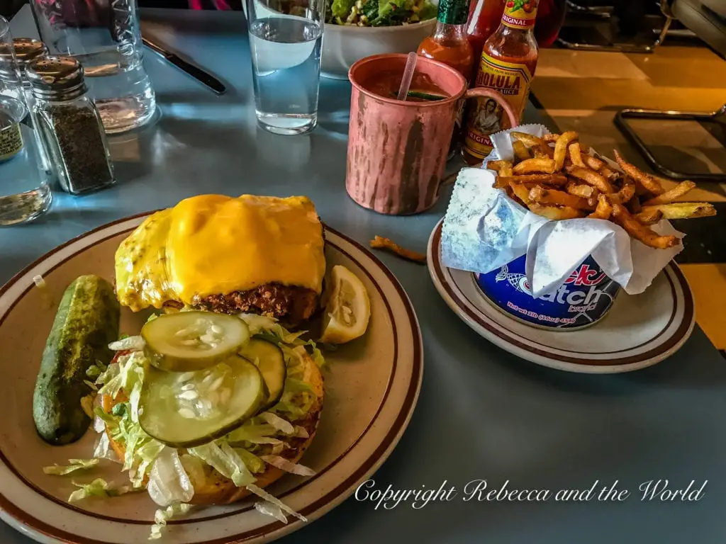 A hearty meal consisting of a cheese-covered burger with lettuce and pickles, a side of fries served in paper, and a copper mug, possibly containing a drink, all on a blue tabletop.