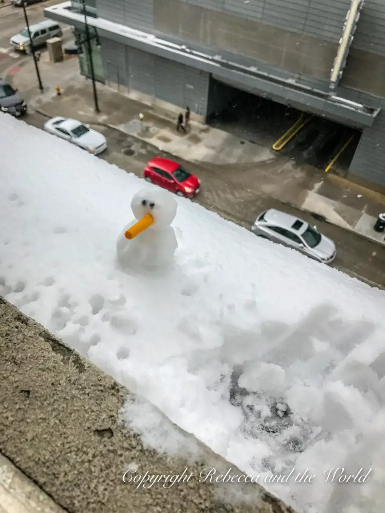 A small, makeshift snowman with an orange nose perched on a snow-covered ledge, overlooking a city street with cars and buildings below.