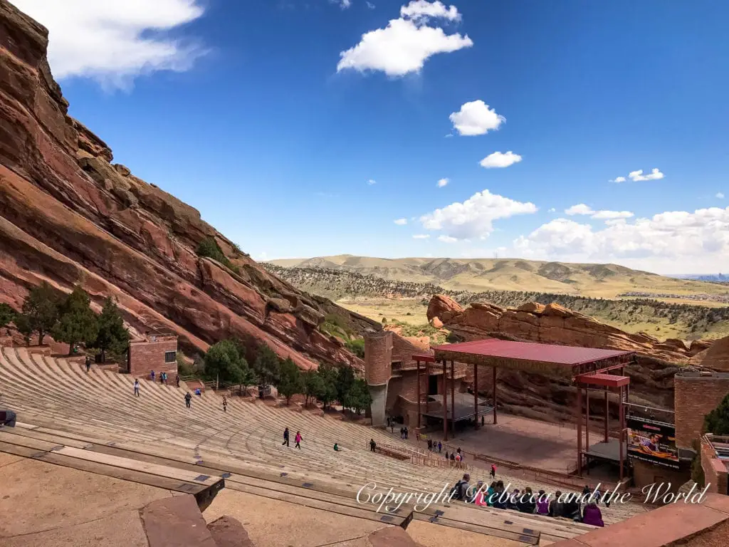 A view from the top of the Red Rocks Amphitheater shows tiered seating down to a stage, with red rocks surrounding the outdoor venue