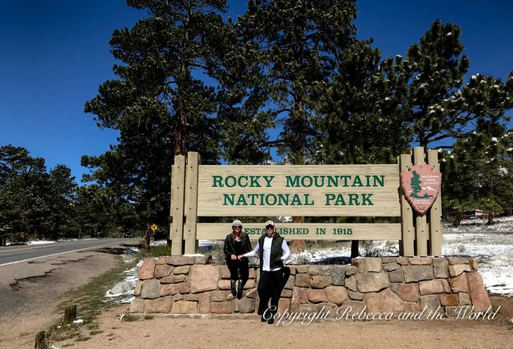 Two people - the author and her husband - posing with smiles in front of the Rocky Mountain National Park entrance sign, surrounded by pine trees with a hint of snow on the ground and clear blue skies above.