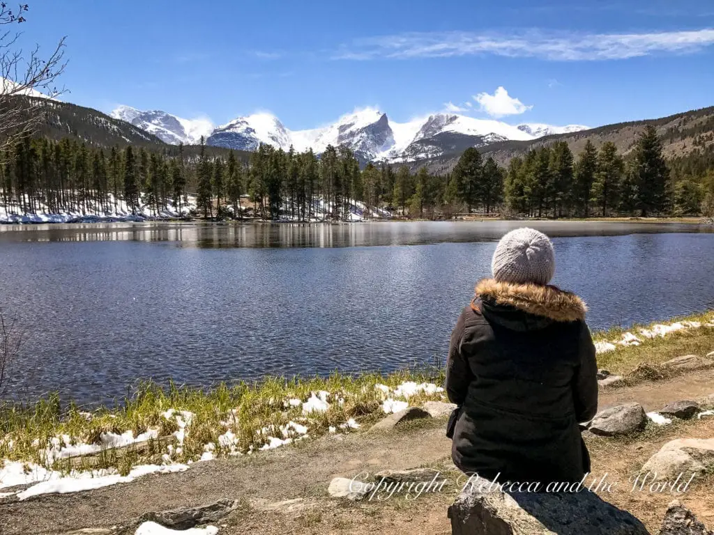 A person - the author of this article - seated on a rock, viewed from behind, gazing out at a tranquil lake surrounded by pine trees with snow-capped mountains in the distance under a clear blue sky.
