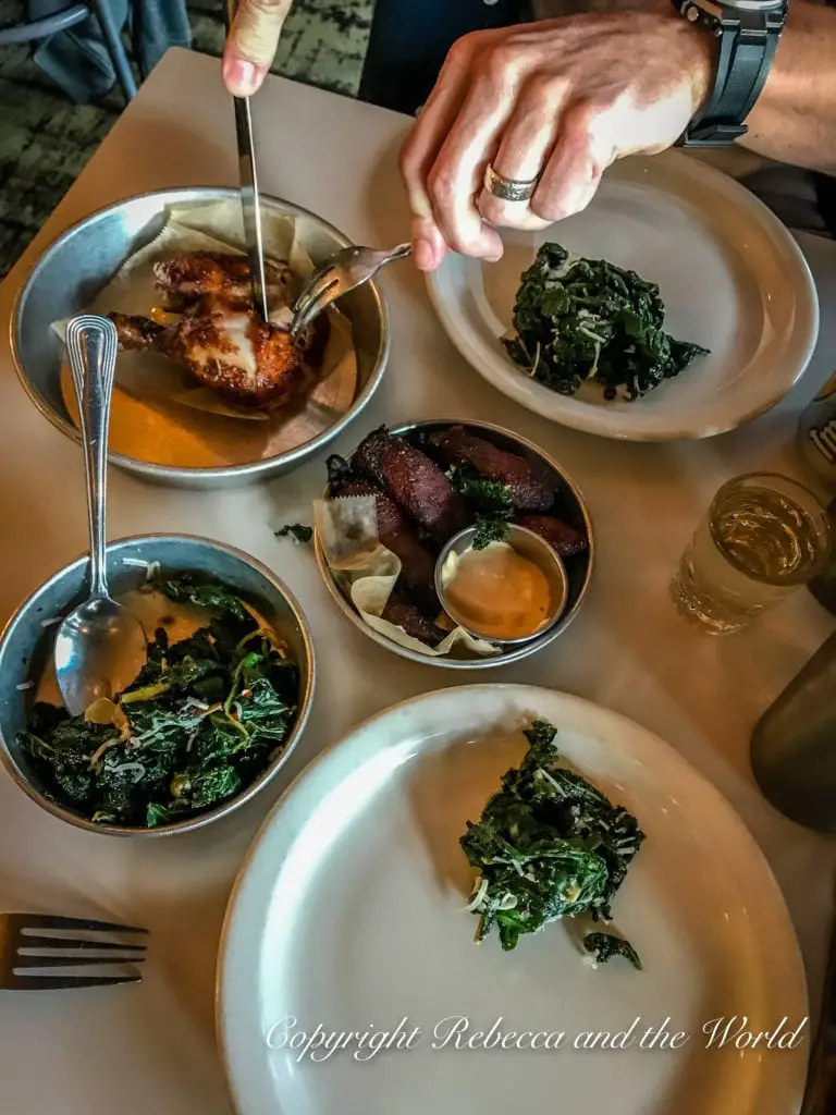 A table with various dishes, including what appears to be roasted chicken, sliced sausage, and sautéed kale. Hands are seen using utensils to serve the chicken.