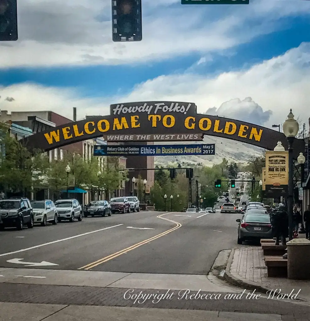 A large welcome sign reading "Howdy Folks! WELCOME TO GOLDEN" in bold letters stretches across a street lined with parked cars and buildings, beneath a partly cloudy sky. Golden CO is a great place to visit on your 3-day Denver winter itinerary.
