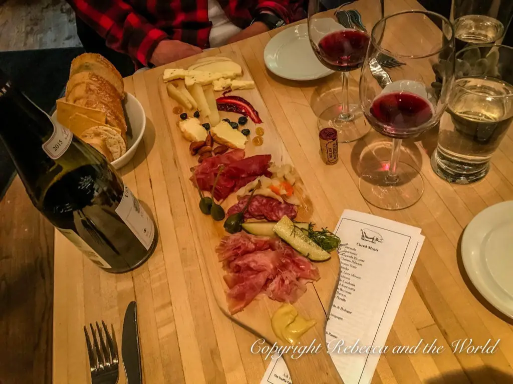 A charcuterie board with an assortment of cheeses, cured meats, and garnishes, accompanied by slices of bread, two glasses of red wine, and a wine bottle on a wooden table.