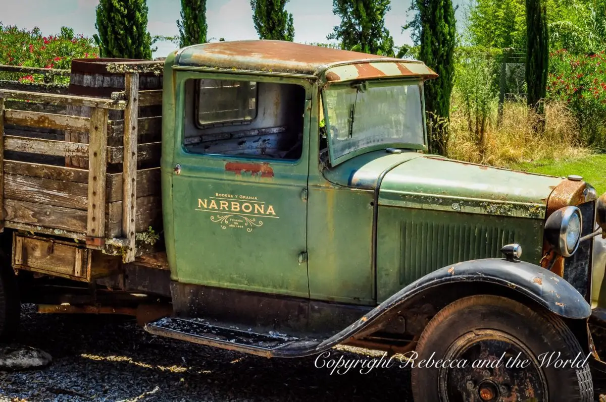 An old green truck with 'Narbona' written on the side is parked beside a vine-covered fence with trees in the background.