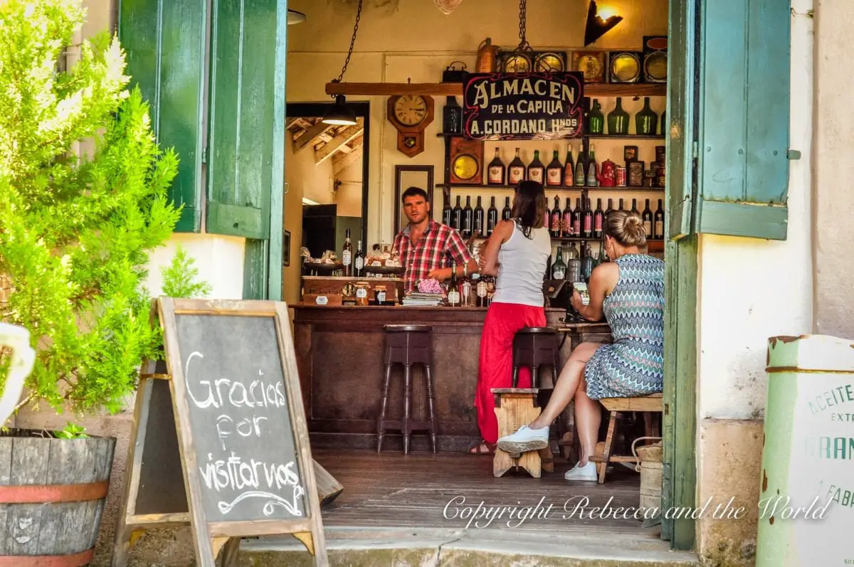 Inside a cozy bar with open green shutters, a bartender stands behind the counter while patrons sit at stools and a table. This is Almacen de la Capilla in Carmelo, Uruguay.