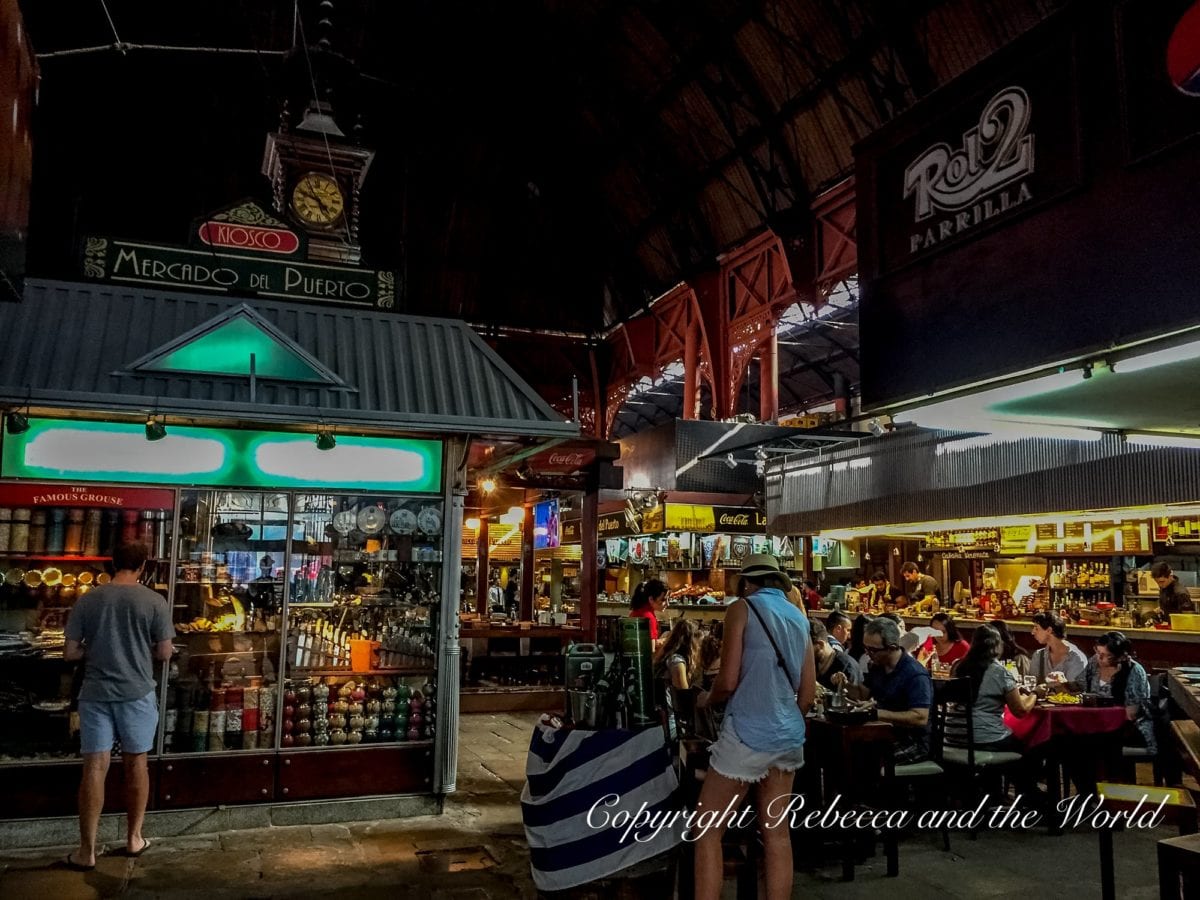 A bustling indoor market with various stalls and eateries. People are dining and walking under the market's high arched ceiling. This is the Mercado Puerto in MOntevideo, Uruguay. Montevideo is a must-visit on your one week in Uruguay itinerary.