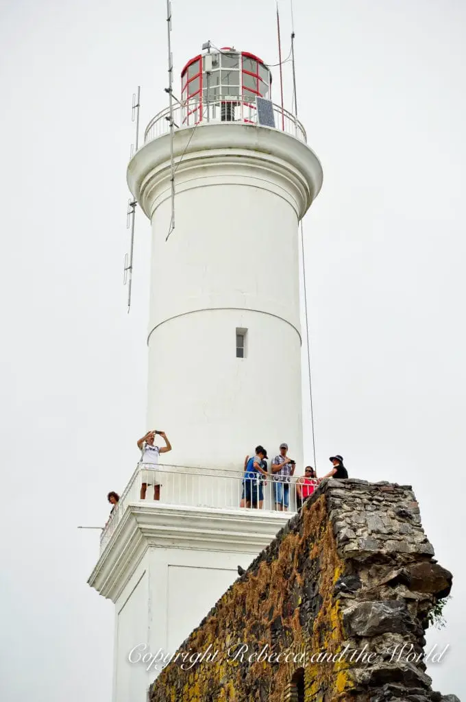 A white lighthouse stands atop a rugged cliff. People gather around the balcony at the top, overlooking the area. This is El Faro, the lighthouse, in Colonia del Sacramento, which should be your first stop on a coastal Uruguay road trip.