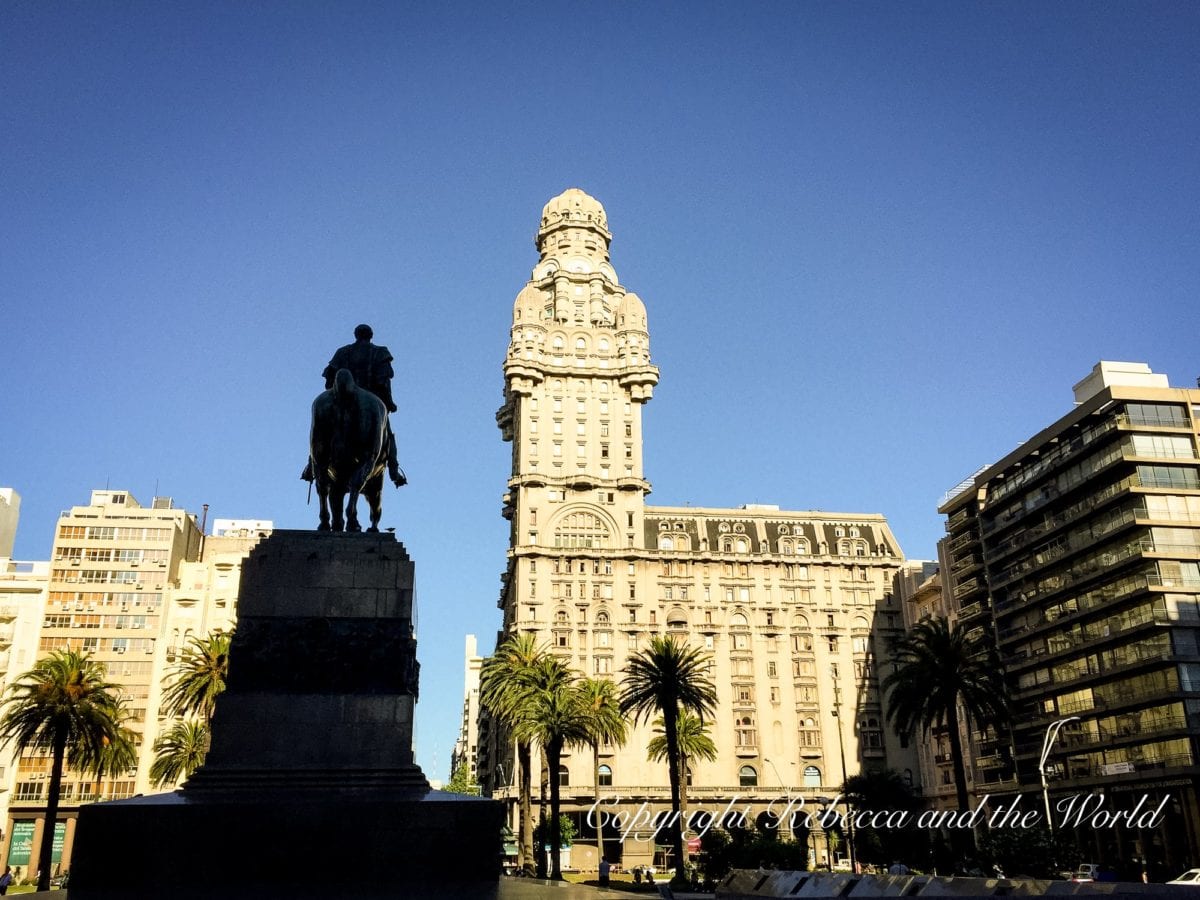 A statue of a historical figure on horseback in front of a tall, ornate building with palm trees and modern buildings around it. This is Plaza Independencia in Montevideo, Uruguay.