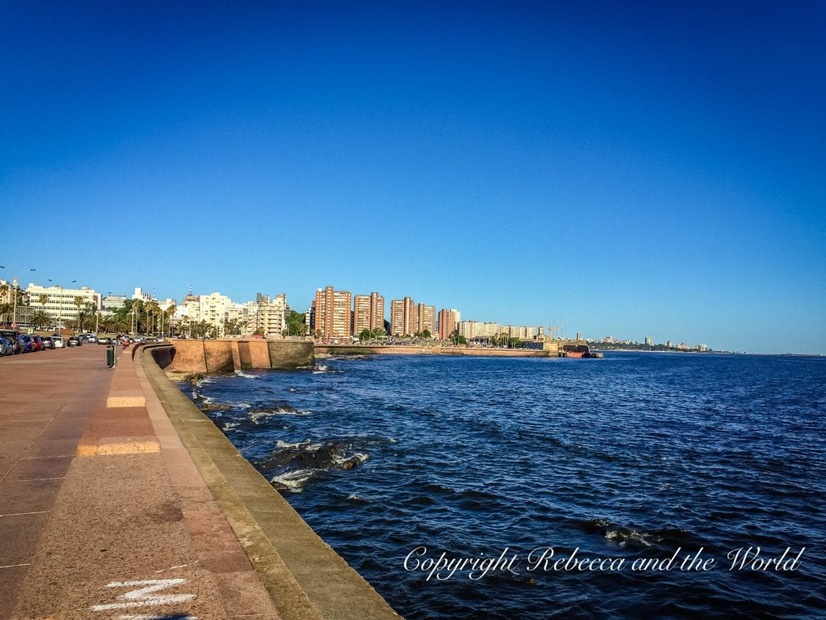 A wide promenade beside a calm blue sea, with a cityscape and clear sky in the background. This lovely spot is La Rambla, one of the best things to do in Montevideo, Uruguay.