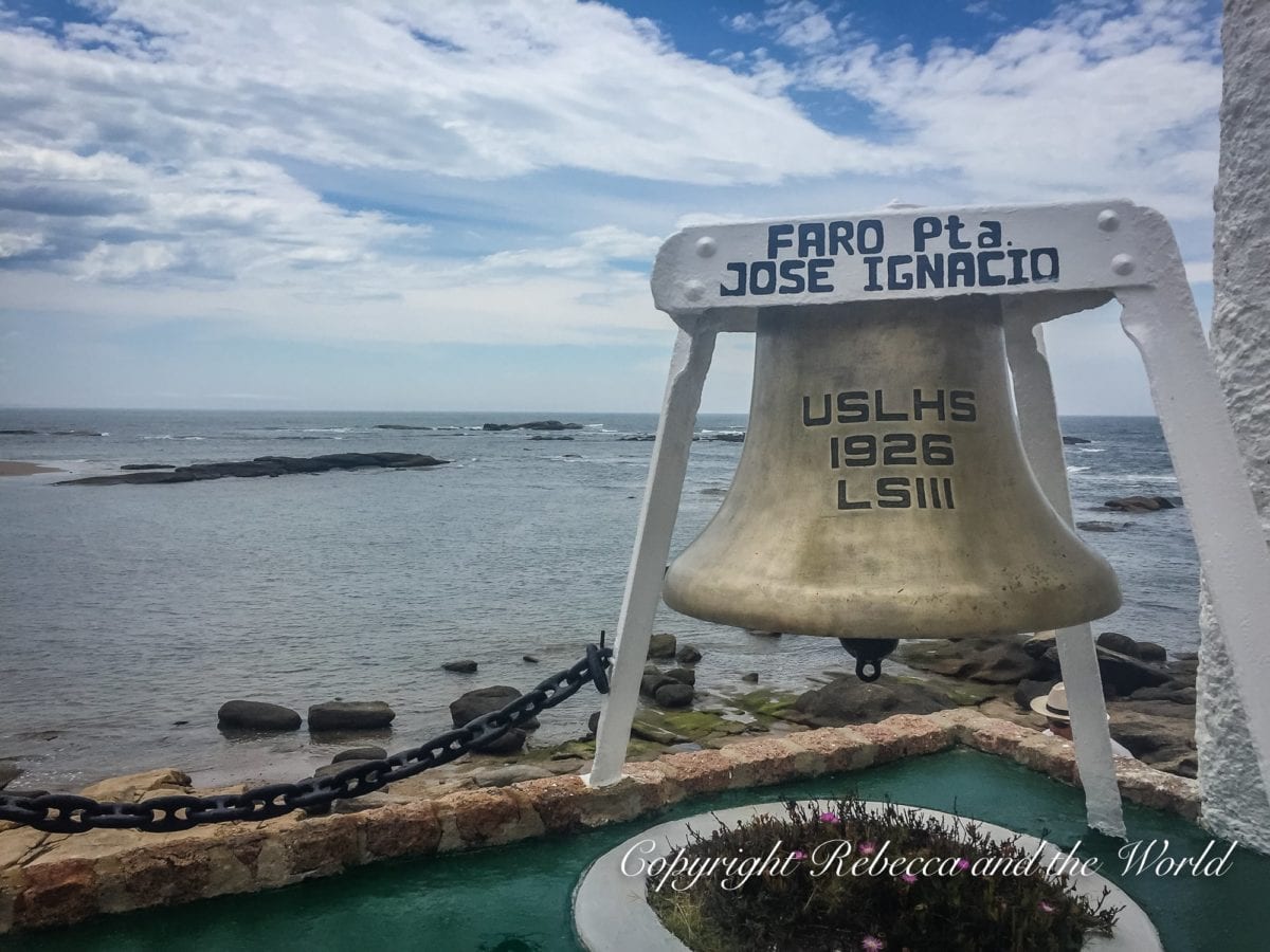 A large, old bell with the inscription "FARO Pta. JOSE IGNACIO USLHS 1926 LSIII" stands before a seascape with rocky outcrops. It's found in Jose Ignacio, a small town in Uruguay.