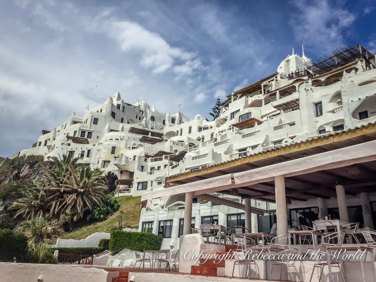 A unique white building with a cascade of irregular windows and terraces perched on a rocky hill, overlooking a beachfront. This building can be found in Punta del Este, Uruguay.