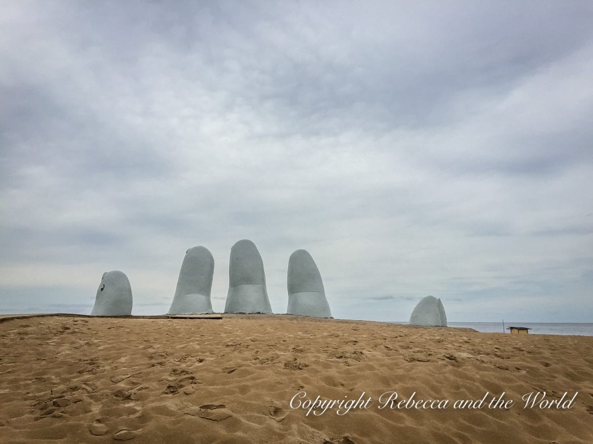 A sandy beach with a sculpture of five oversized fingers partially buried, reaching up to the sky, under a cloudy backdrop. This is La Mano en La Arena sculpture in Punta del Este, Uruguay.