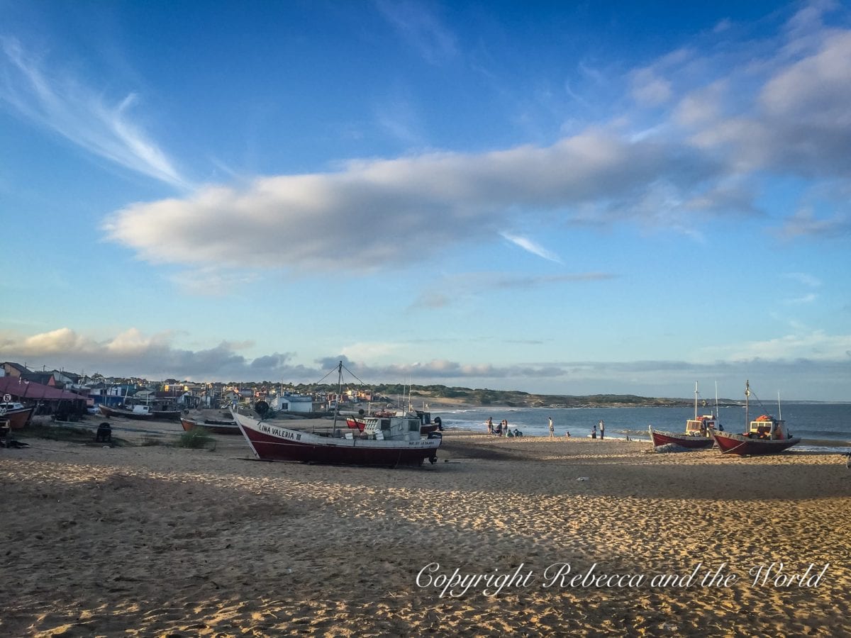 Fishing boats on a sandy shore with a backdrop of a coastal town and clear skies with wispy clouds at dusk. This is Punta del Diablo, on the coast of Uruguay, a great stop on your Uruguay itinerary.