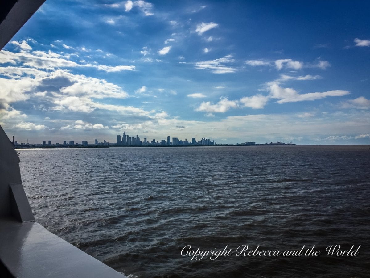 A view from a boat showing a distant city skyline across the water, under a sky with scattered clouds. This is the view of Buenos Aires from the ferry.