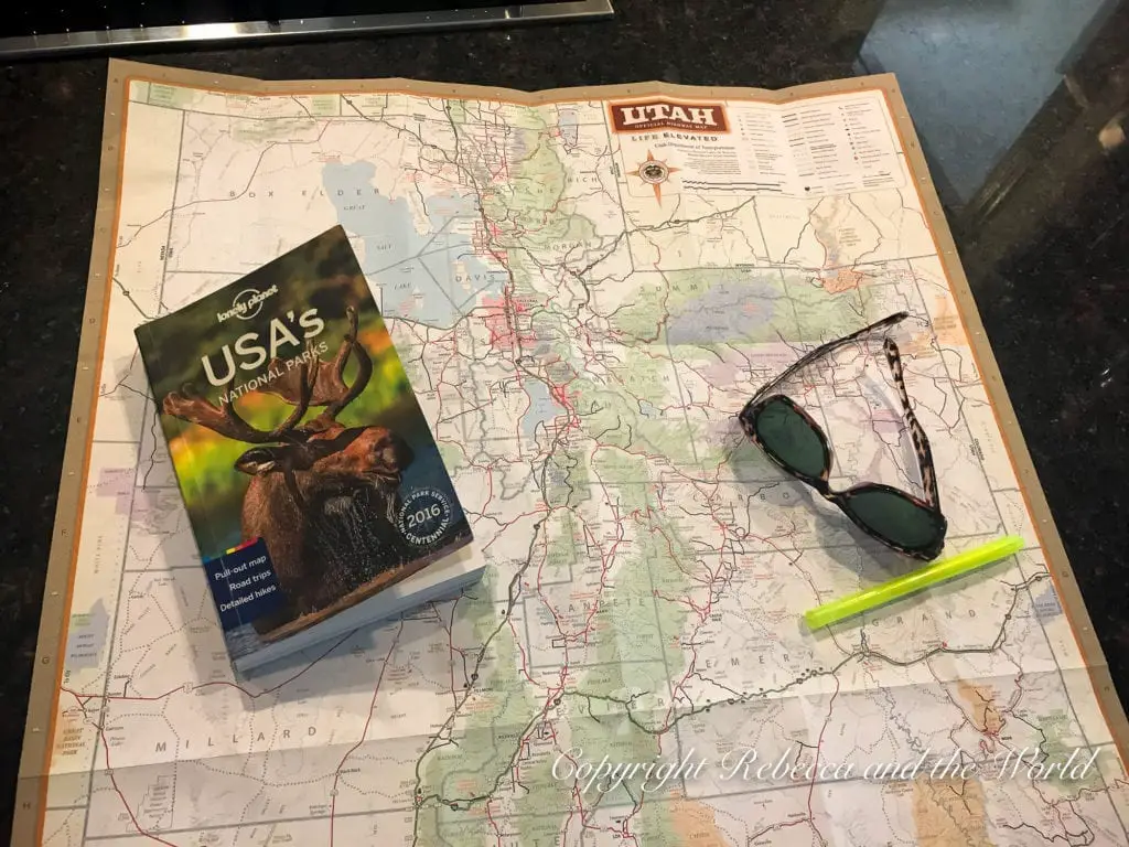 A large, unfolded map of Utah spread out on a dark surface with a guidebook titled "USA's National Parks" and a pair of sunglasses and a yellow highlighter resting on top.