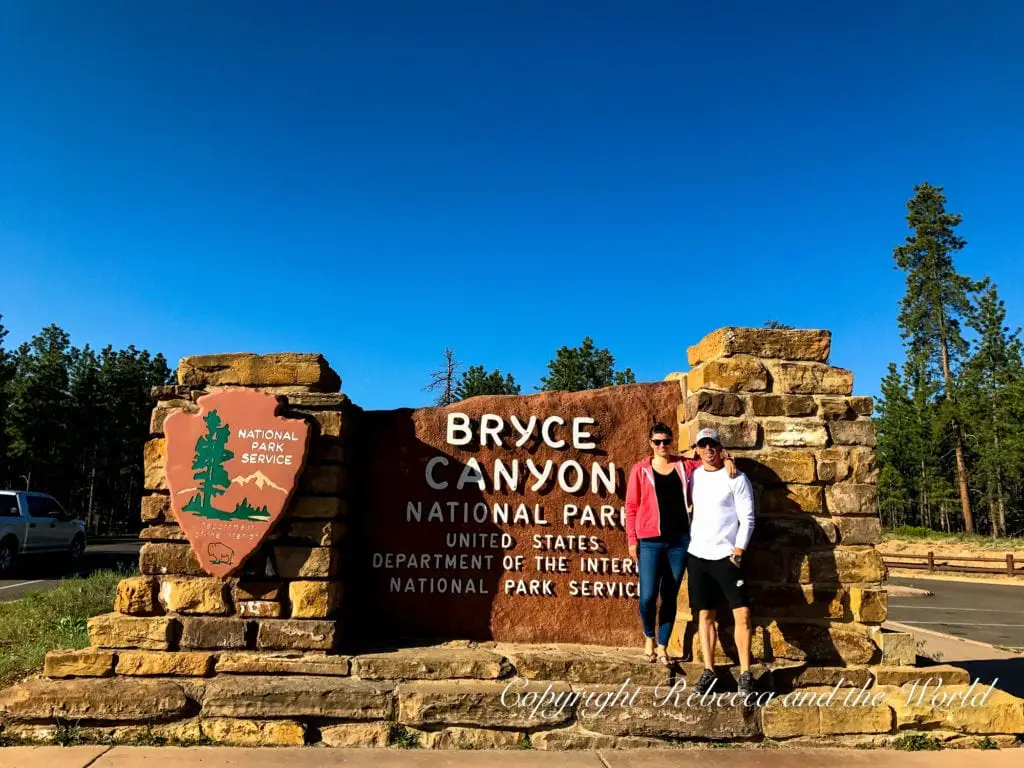 Two people - the author of this article and her husband - stand in front of the Bryce Canyon National Park sign, composed of stone and wood, under a clear blue sky. Trees are visible in the background.