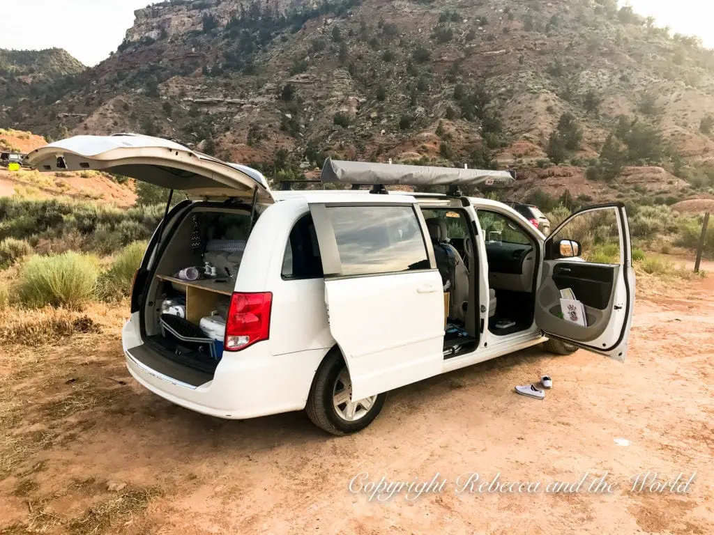 A white minivan with its rear and side doors open, revealing camping gear inside, parked on a dirt area with scrub vegetation, with red rock formations and a hill in the background under a clear sky. The campervan we travelled with on our Utah national parks road trip.