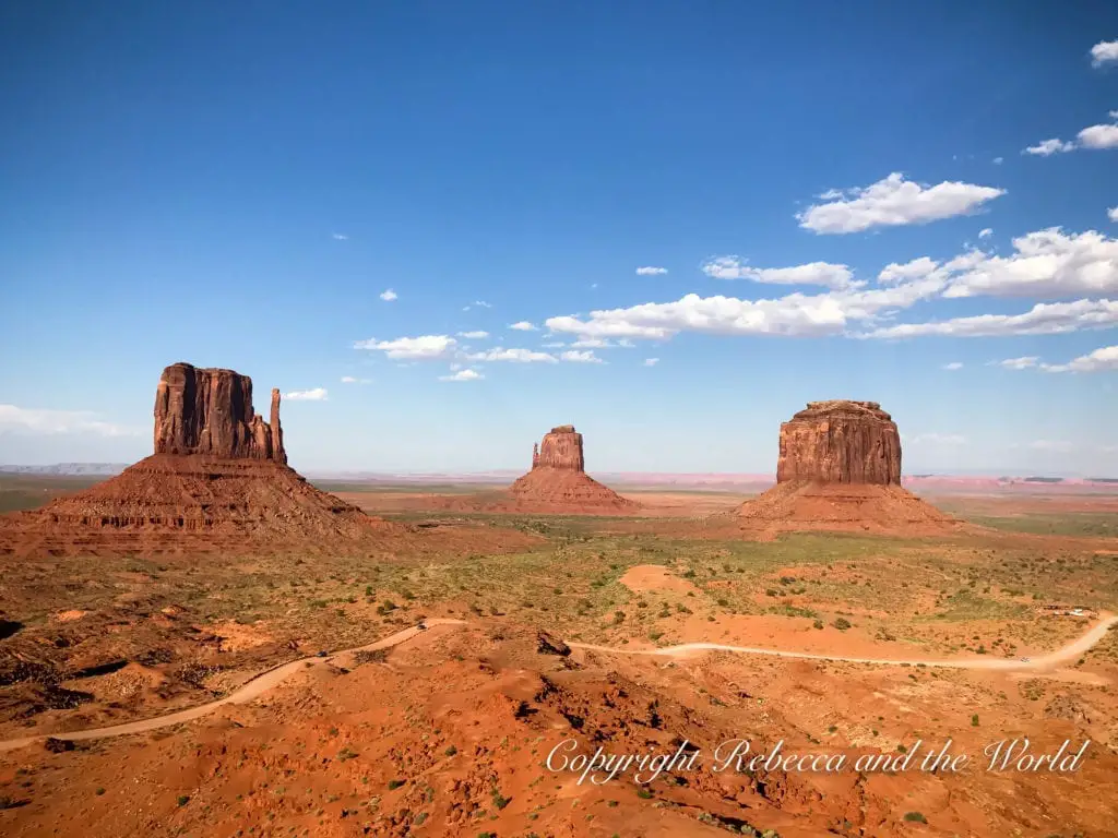 The iconic Mittens and Merrick Butte in Monument Valley under a bright blue sky with few clouds. The formations stand prominently on a flat, desert landscape.