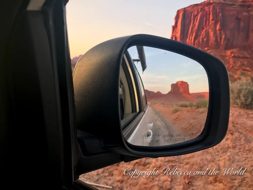 A vehicle's side mirror reflects a view of a red rock formation in the desert during sunset, with the text "Objects in mirror are closer than they appear" visible. This was taken in Monument Valley in Arizona.