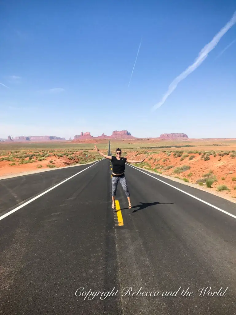 A woman - the author of this article - stands on the centerline of a straight asphalt road stretching into the distance with Monument Valley's rock formations visible on the horizon under a clear blue sky.