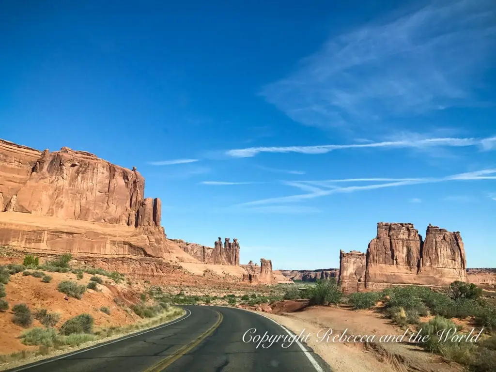 A road leading through Arches National Park with tall red sandstone formations on either side under a sky with wispy clouds.
