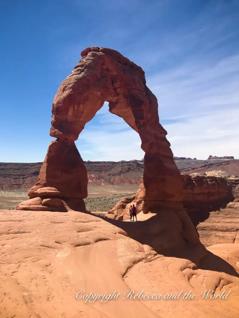The Delicate Arch in Arches National Park, a standalone natural arch with a clear blue sky in the background, with two people - the author of this article and her husband - standing underneath for scale.