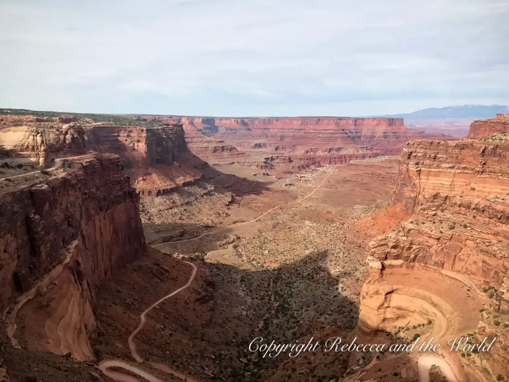 A view of Shafer Canyon in Canyonlands National Park, with a winding dirt road descending into the valley, flanked by steep red cliffs and vast desert plateaus extending into the distance.