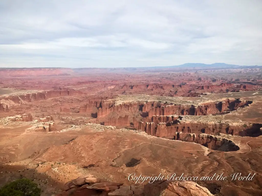 A sweeping vista of the Island in the Sky district of Canyonlands National Park, featuring expansive flat-topped mesas with deep canyons, in layers of red and orange, under a hazy sky.