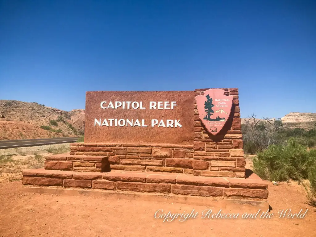 The entrance sign for Capitol Reef National Park, composed of red brick with a carved wooden park service emblem, set against a desert landscape and clear blue sky.