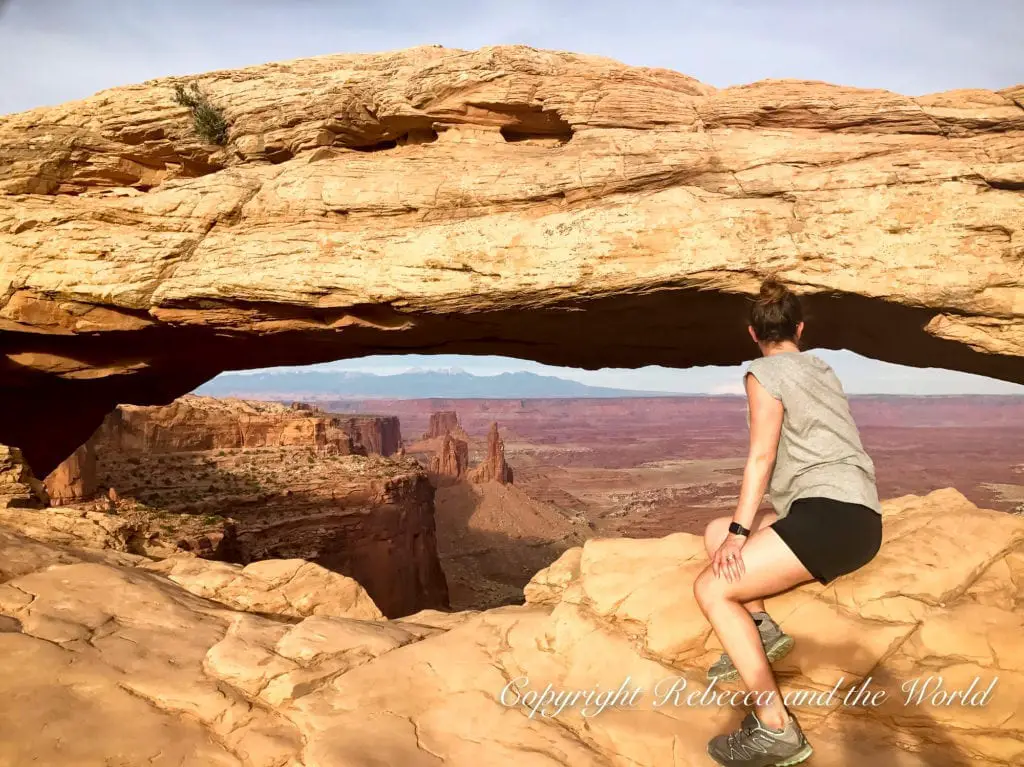 A woman - the author of this article - sits under Mesa Arch, a natural stone arch in Canyonlands National Park, framing a distant view of the park's canyons and plateaus under a clear sky.