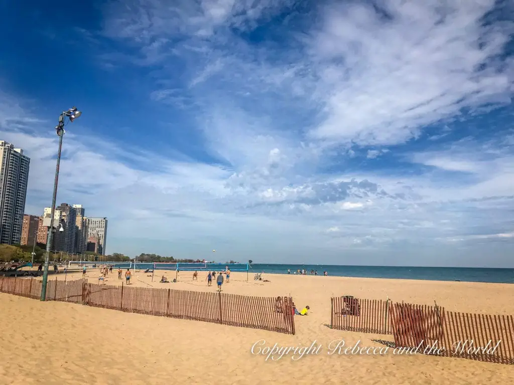 A sandy beach with people playing volleyball and relaxing near the Lake Michigan shore, with Chicago's high-rise buildings in the distance.