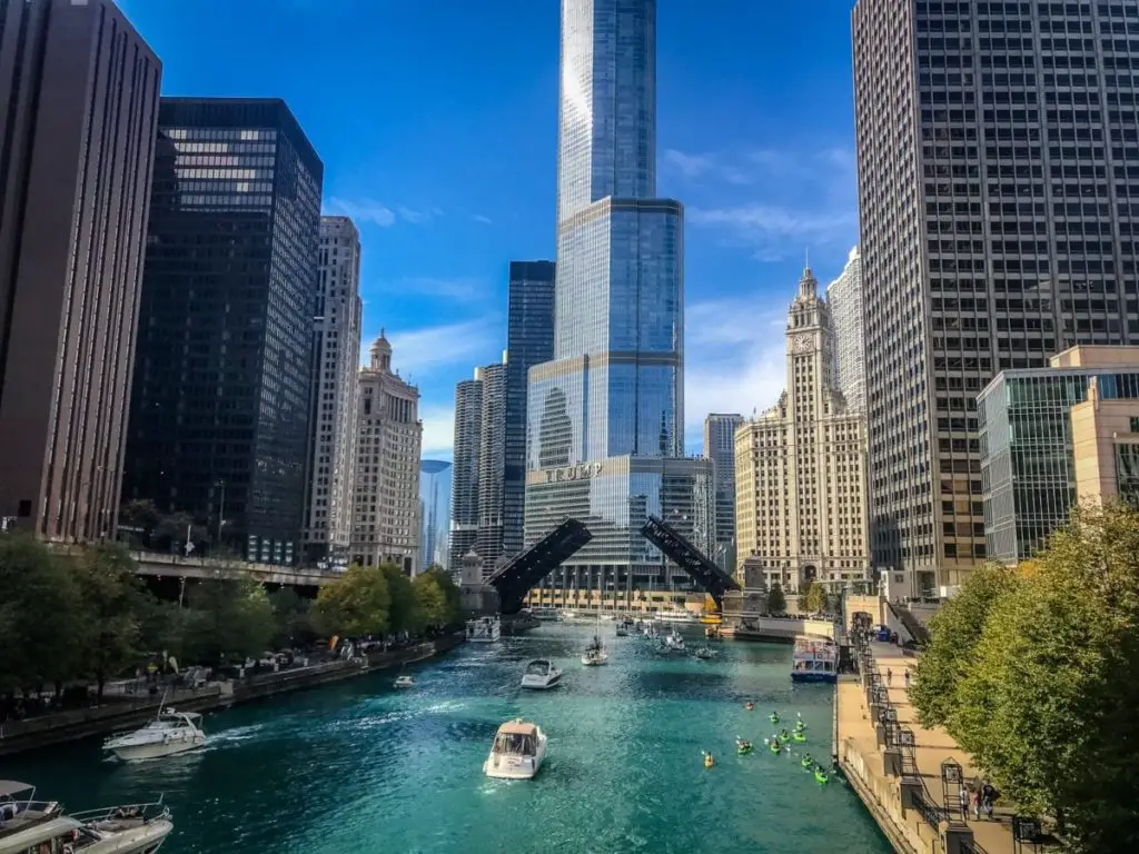 The Chicago River flanked by towering skyscrapers and a clear blue sky. Boats navigate the water as pedestrians walk along the riverwalk.