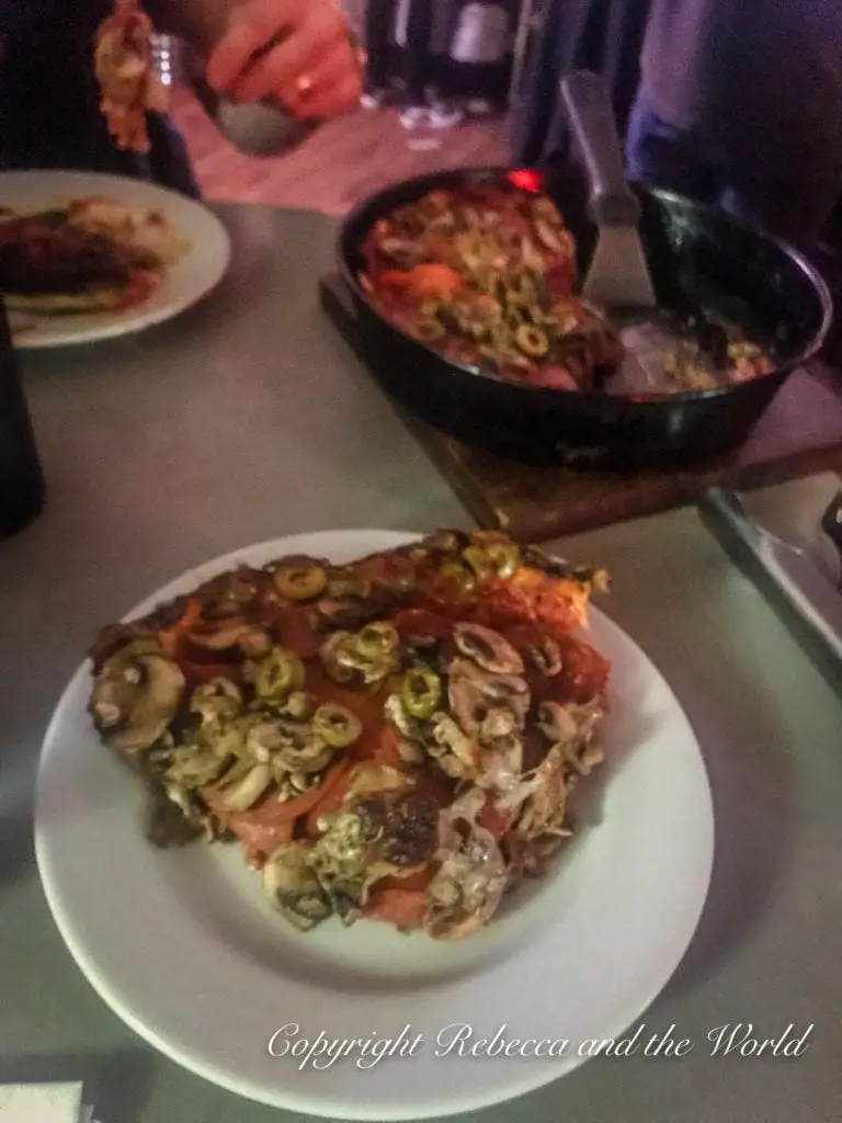 A close-up view of a Chicago-style deep-dish pizza on a white plate, with toppings of olives, mushrooms, and tomatoes, accompanied by a cast iron skillet with more pizza in the background.