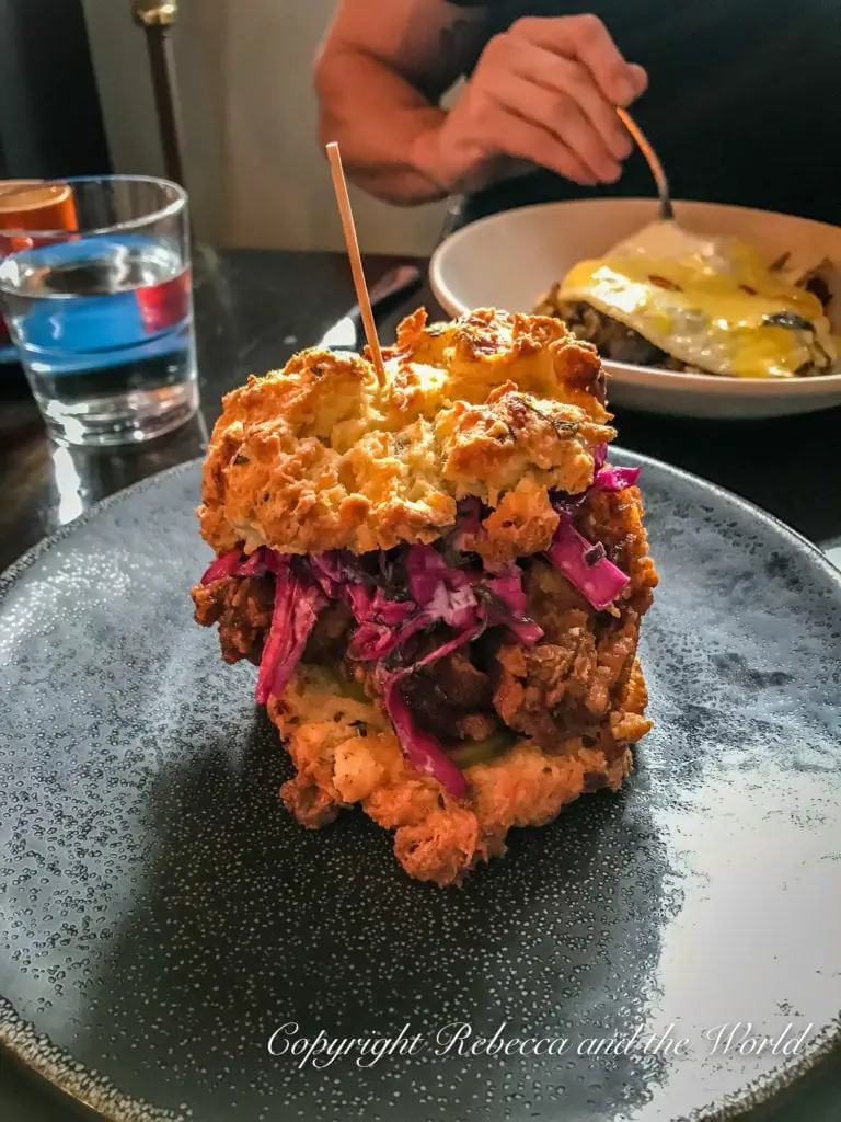 A gourmet sandwich featuring crispy fried chicken and red cabbage slaw between two flaky biscuits, skewered with a toothpick, presented on a dark, textured plate. You can fit in a lot of meals in 48 hours in Chicago!
