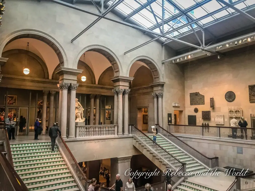 Interior of the Art Institute of Chicago, featuring a grand staircase, classical architecture, and visitors admiring the artwork.