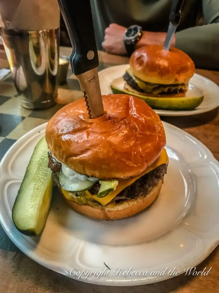 A cheeseburger with multiple patties, cheese, pickles, and onions, served on a glossy brioche bun with a steak knife stabbed through the center for support, next to a pickle spear. Au Cheval's burgers are one of the most famous things to eat in Chicago.