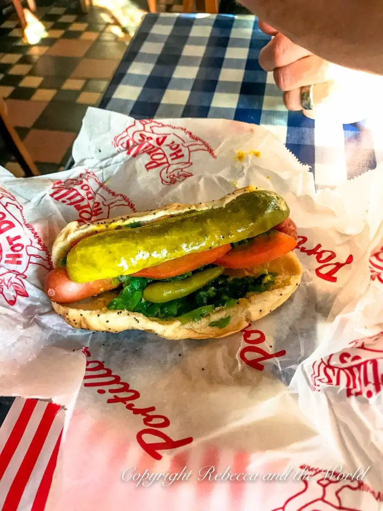 A classic Chicago-style hot dog on a poppy seed bun with mustard, relish, onions, tomatoes, a pickle spear, sport peppers, and a dash of celery salt, wrapped in paper.