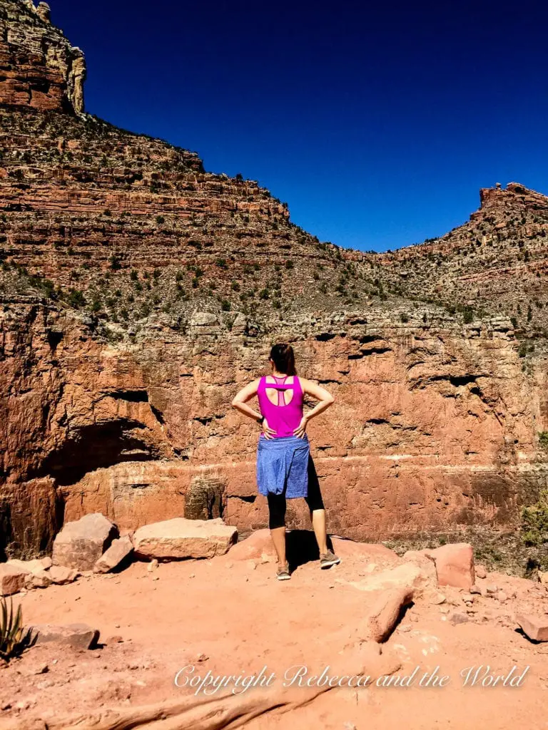 A woman - the author of this article - stands with her back to the camera, looking out over the Grand Canyon. She wears a pink tank top, black leggings and a blue sweater around her waist. The canyon's vastness spreads out before her, with steep cliffs on either side.