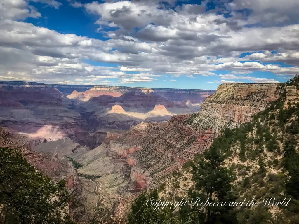 A view of the Grand Canyon under a cloudy sky, with sunlight spotlighting certain areas. The canyon layers display various shades of red, brown, and green, with a winding river visible in the distance.