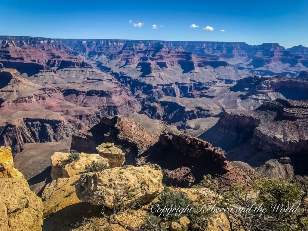 Overhead view of the Grand Canyon with a clear blue sky. The rugged landscape features prominent rock formations and deep valleys, all with distinct layers of colored rock strata.