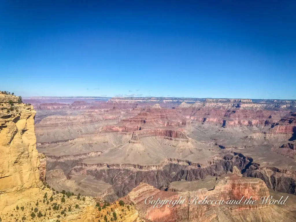 Grand Canyon vista with a vast, open sky. The canyon walls stretch into the distance, displaying a multitude of red and brown hues with green vegetation dotting the landscape.