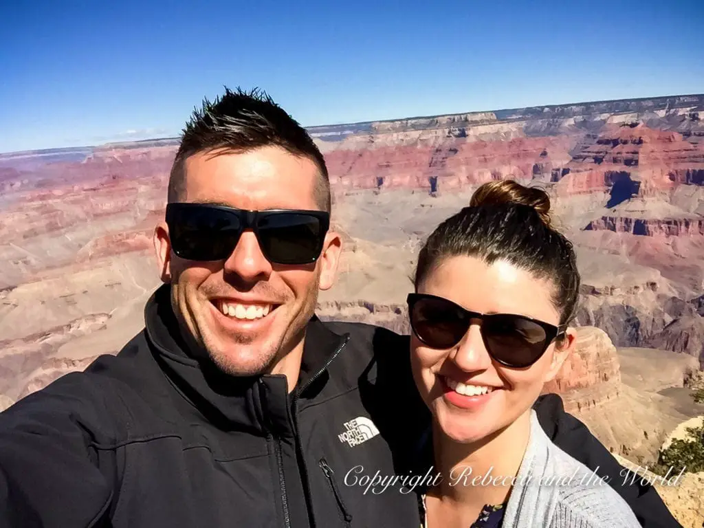 A smiling man and woman - the author of this article and her husband - taking a selfie with the Grand Canyon in the background. Both are wearing sunglasses; the man has short hair and a black jacket, while the woman has her hair in a bun and is wearing a grey top. The vast canyon behind them is a tapestry of red and brown layers under a clear blue sky.